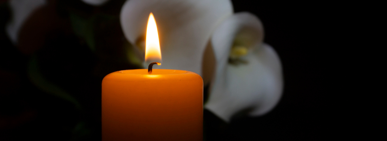 A close up of an orange candle and flame and lily flowers on a dark background.