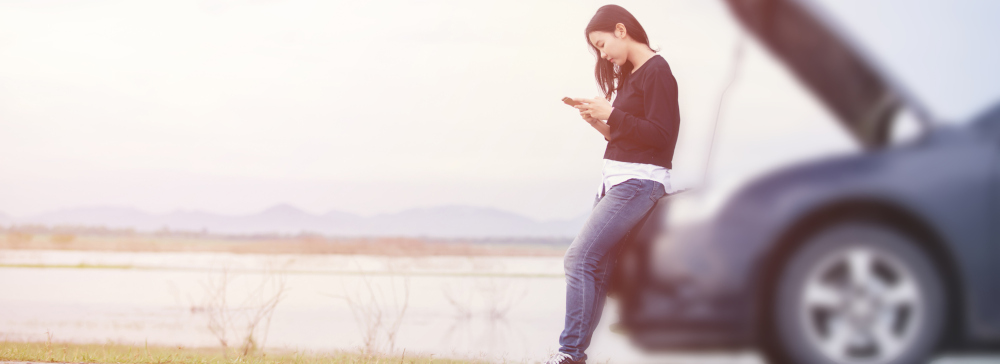 A woman looking at her phone in front of her damaged car after a car accident