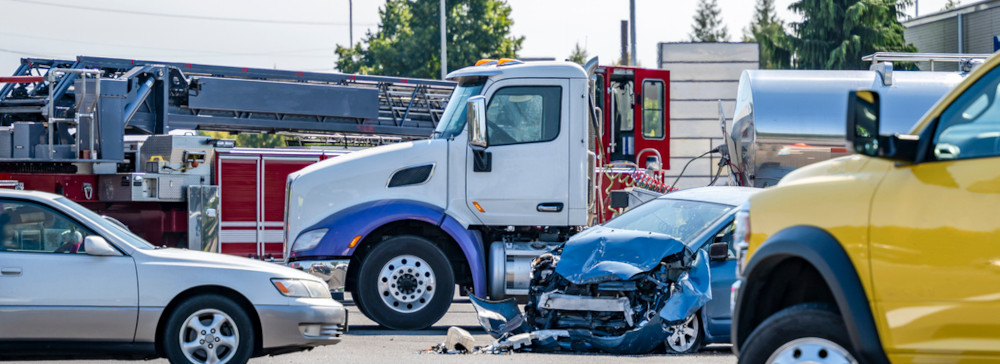 An intersection after a commercial vehicle accident between a blue car and a white 18 wheeler, with a fire truck in the background