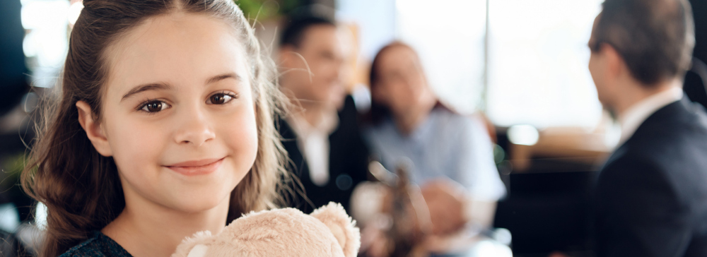 A young girl stands with a teddy bear and smiles at the camera while her parents are in a personal injury legal consultation.
