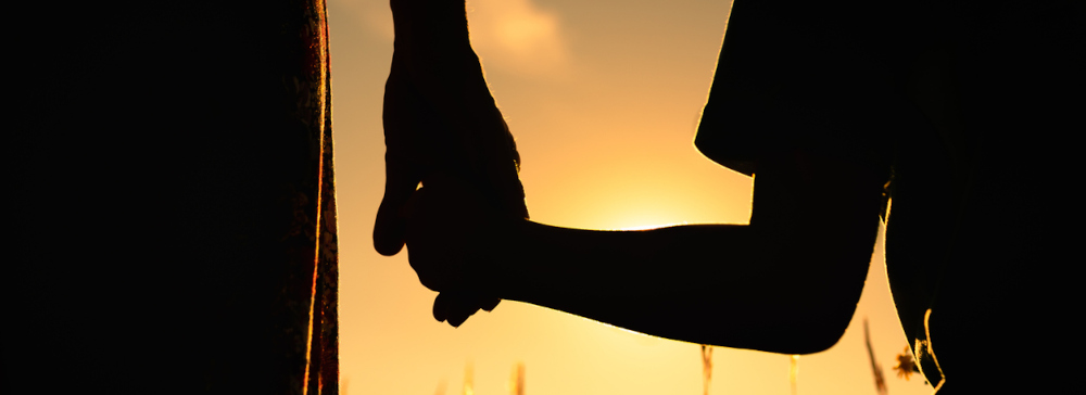 Silhouette of mother and child holding hands in front of a sunset.